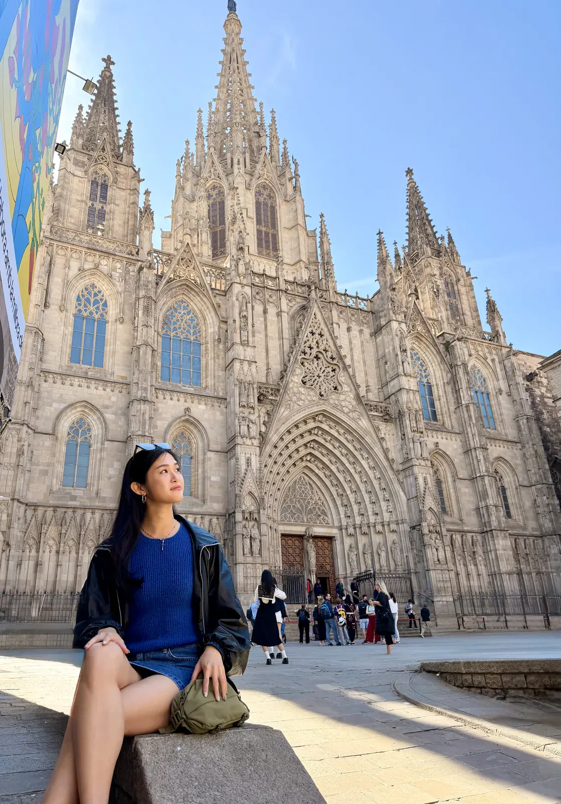 A woman sitting in the square below the Gothic spires and carved doorway of the Cathedral of Barcelona.