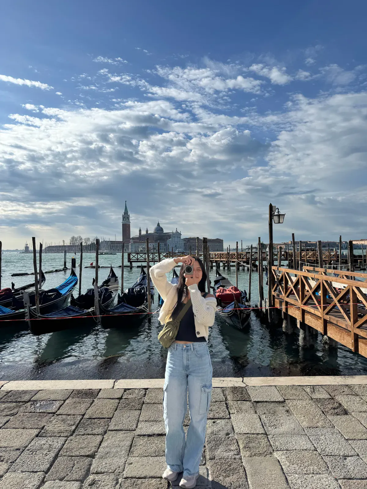 A traveler on the Piazza San Marco waterfront holding up a camera, with gondolas moored behind and the tower and dome of San Giorgio Maggiore across the water