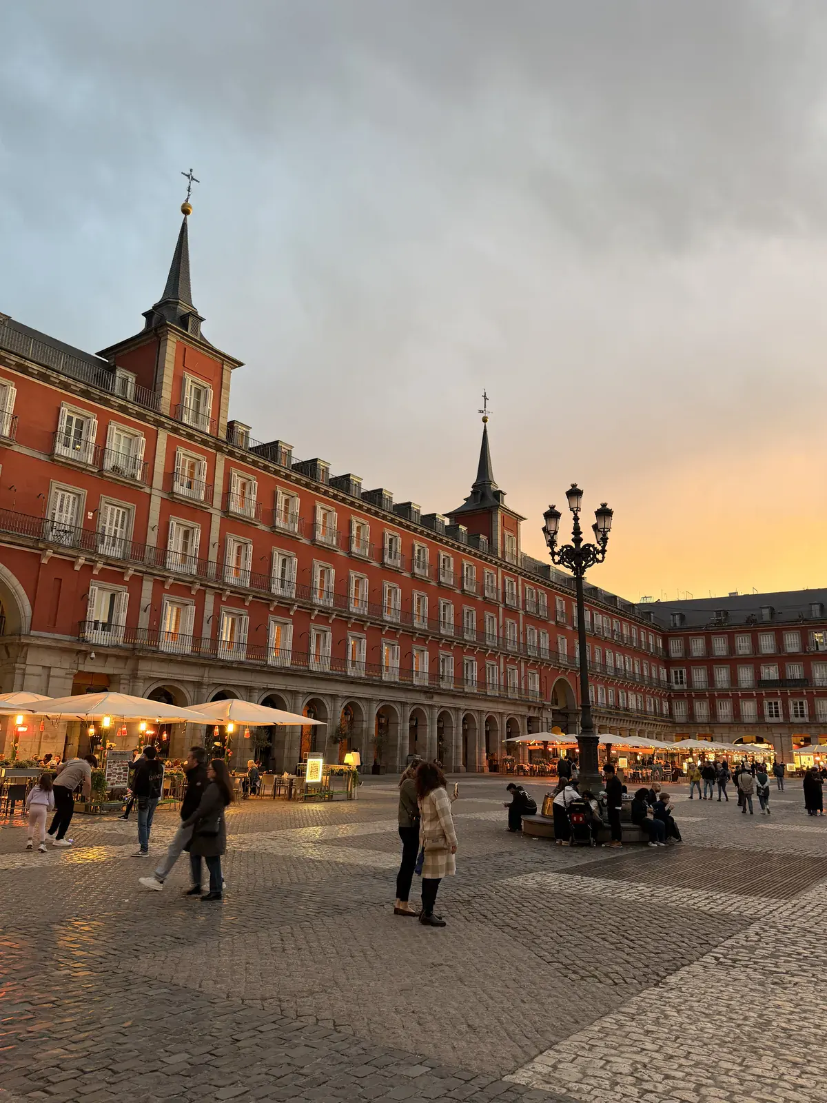 Plaza Mayor's red-brick facades and arcades in Madrid at sunset, with people crossing the cobbled square.