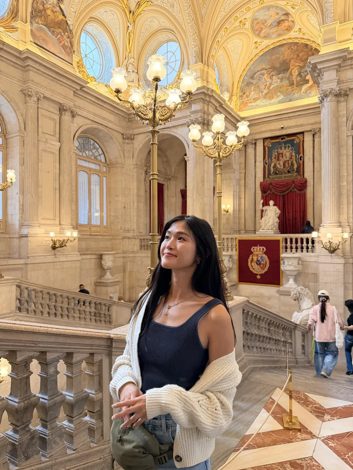 A woman on the Royal Palace of Madrid's main staircase, below frescoed vaults and gilded chandeliers.
