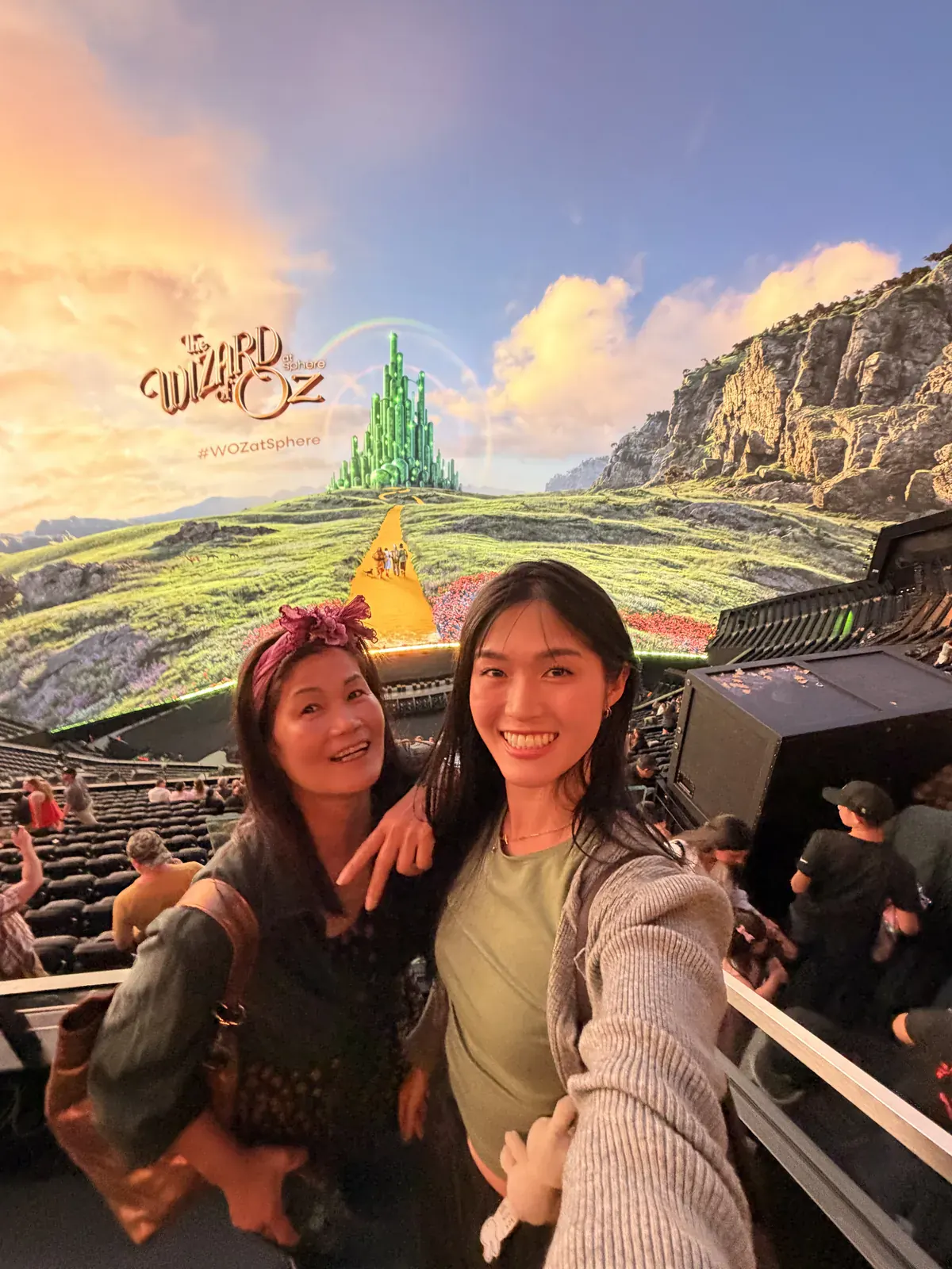 Two visitors in the seating bowl at Sphere in Las Vegas, with the Emerald City and the yellow brick road filling the screen behind them